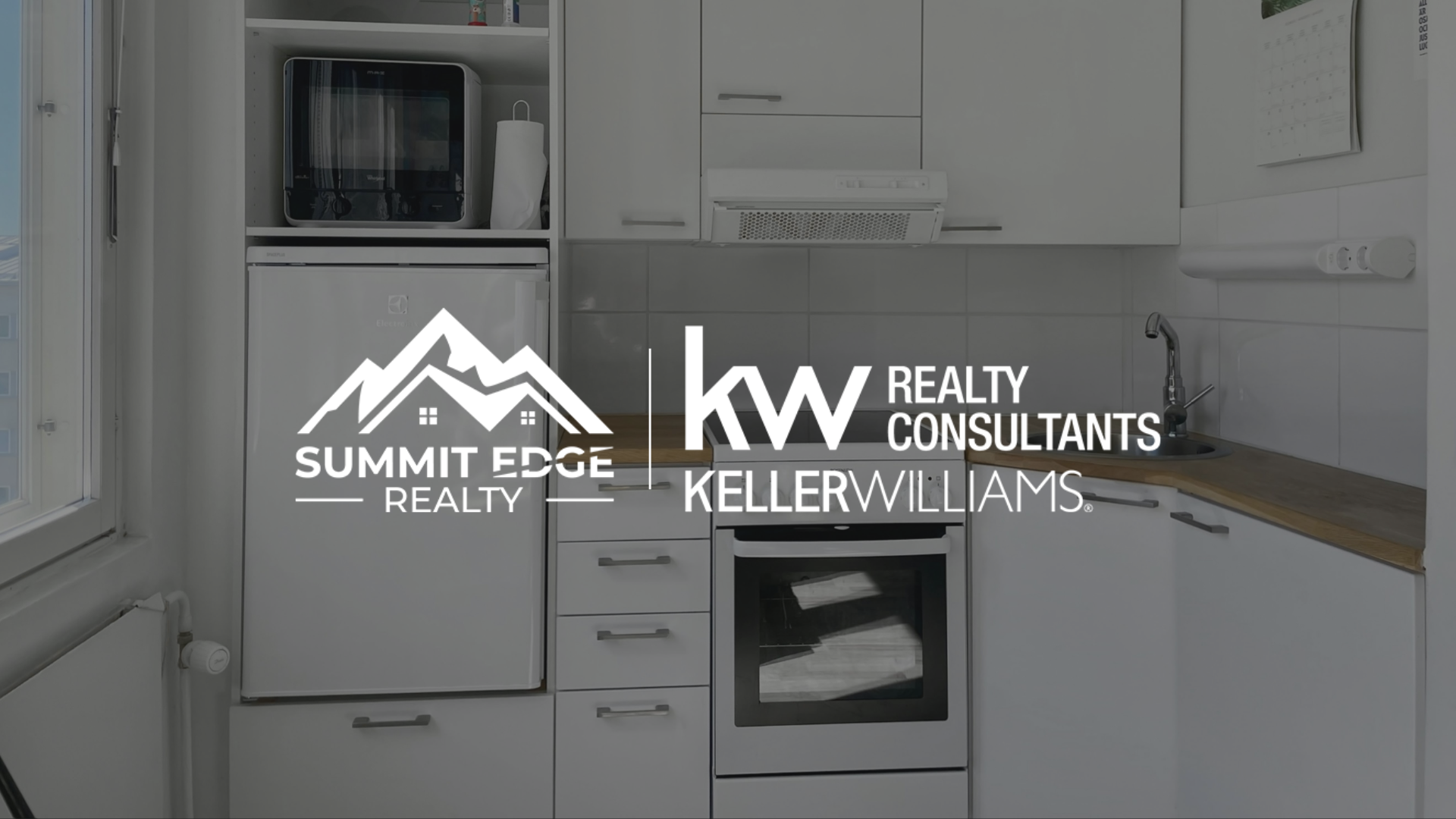 A tiny home kitchen interior with white cabinets, a built-in oven, a microwave, a sink, and a wooden countertop, enhanced by a large window with natural light, overlaid with the Summit Edge Realty and KW Realty Consultants Keller Williams logo in white."