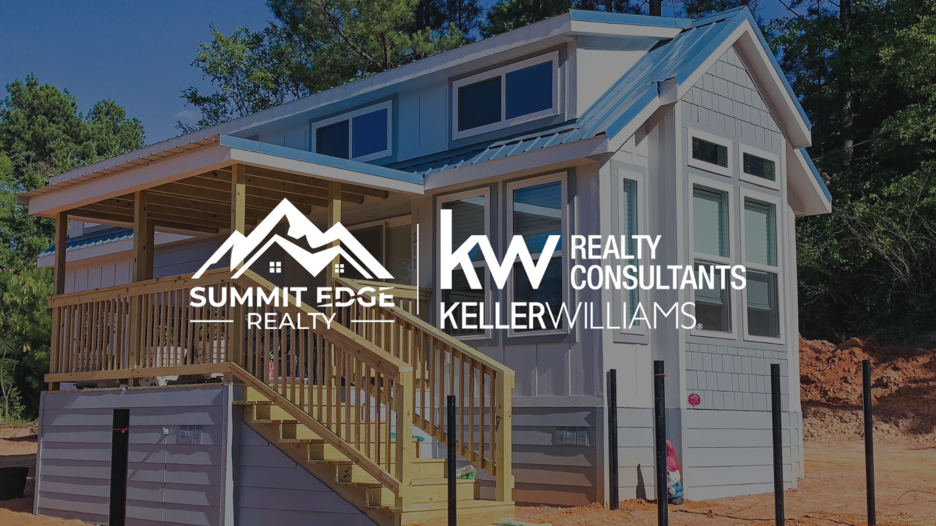 A Clark County, IN, tiny residence with a gray exterior, large windows, and a blue metal roof, including a wooden deck with stairs, situated on a dirt construction site surrounded by pine trees, overlaid with the Summit Edge Realty and KW Realty Consultants Keller Williams logo in white.