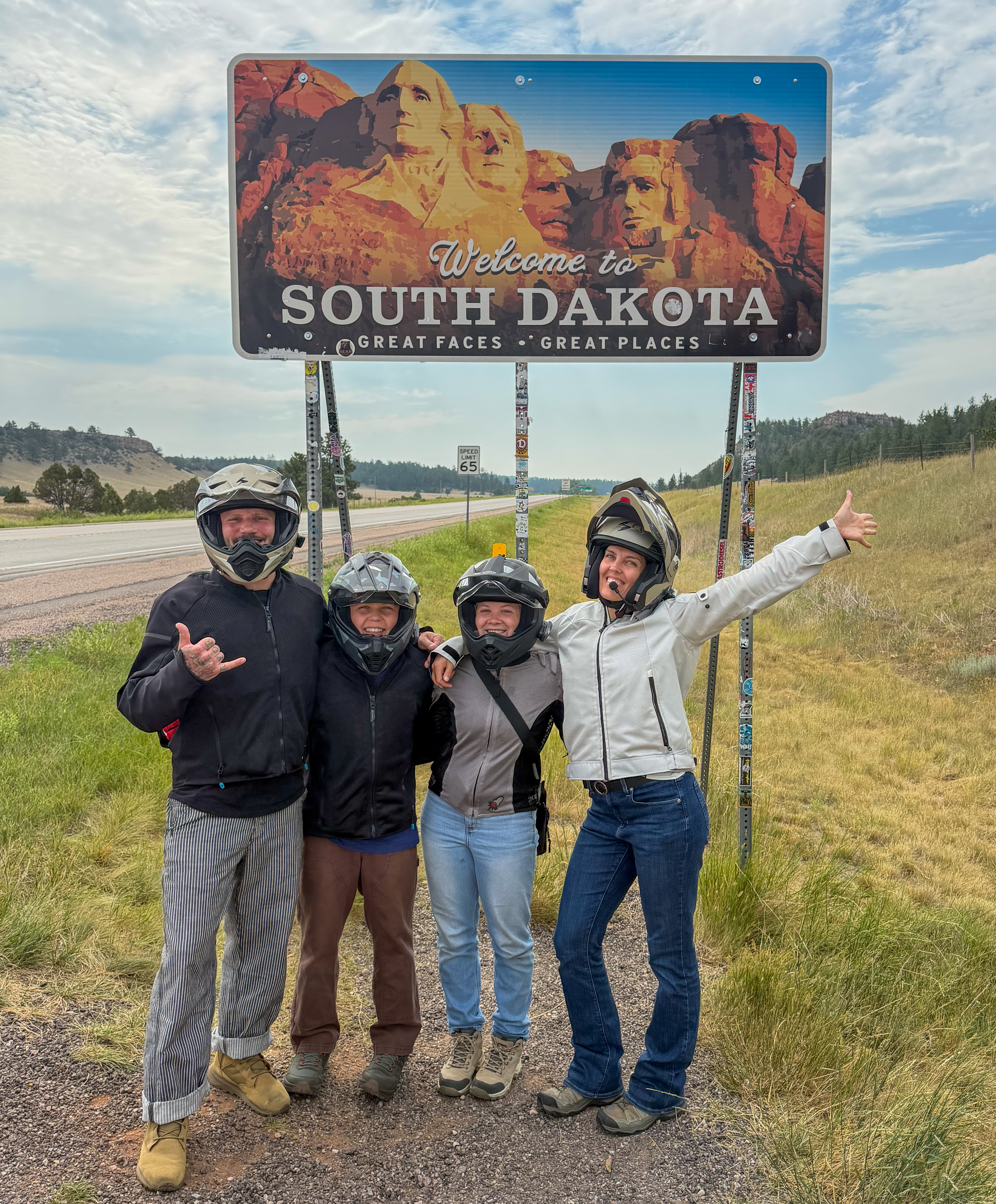 Mathews family stands below the South Dakota state line sign on the side of the highway, wearing motorcycle helmets