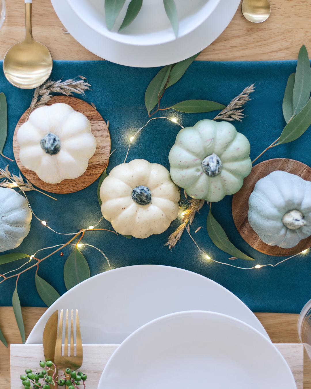 Elegant holiday table setting with mini pumpkins, eucalyptus leaves, and warm string lights.