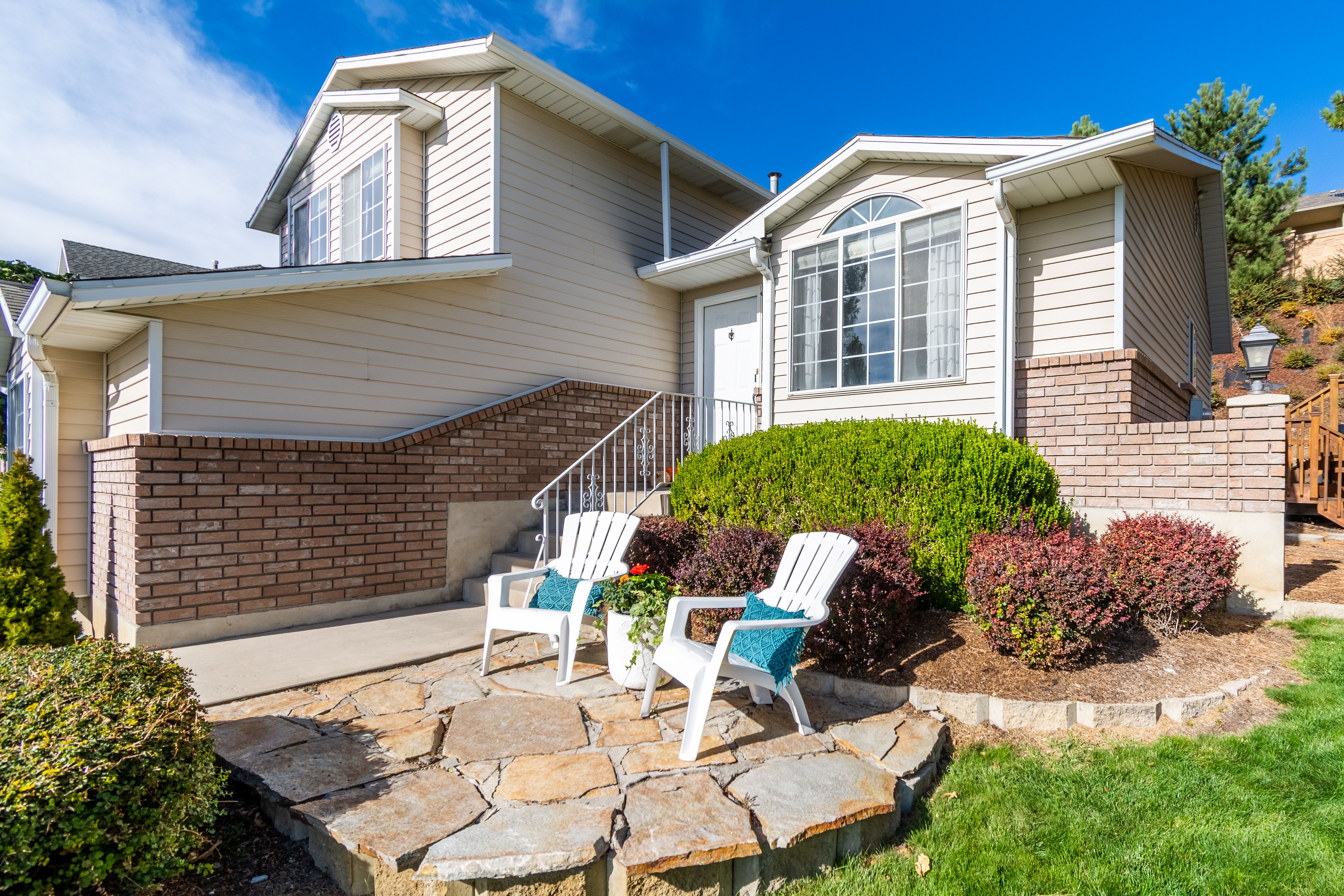 Front exterior of a beige two-story home with brick accents, large picture window, landscaped front yard, and stone patio seating area.