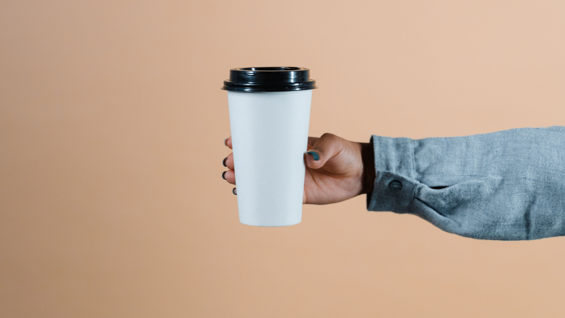 A hand holding a plain white takeaway coffee cup with a black lid against a warm beige background, creating a clean and minimalist coffee-themed image with ample copy space.