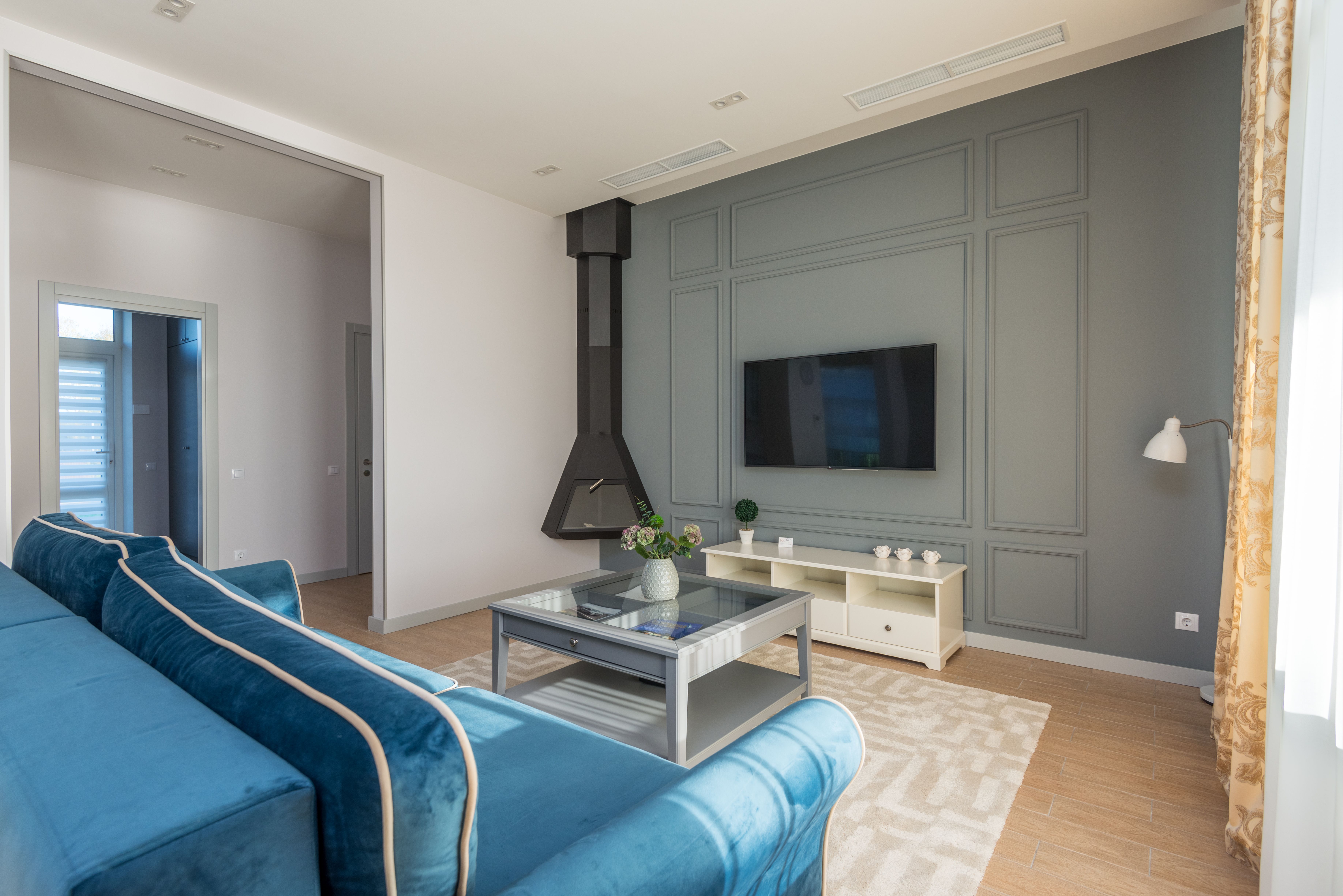 Modern living room with a blue sofa, gray accent wall, mounted TV, contemporary fireplace, and large windows creating a bright, inviting space.
