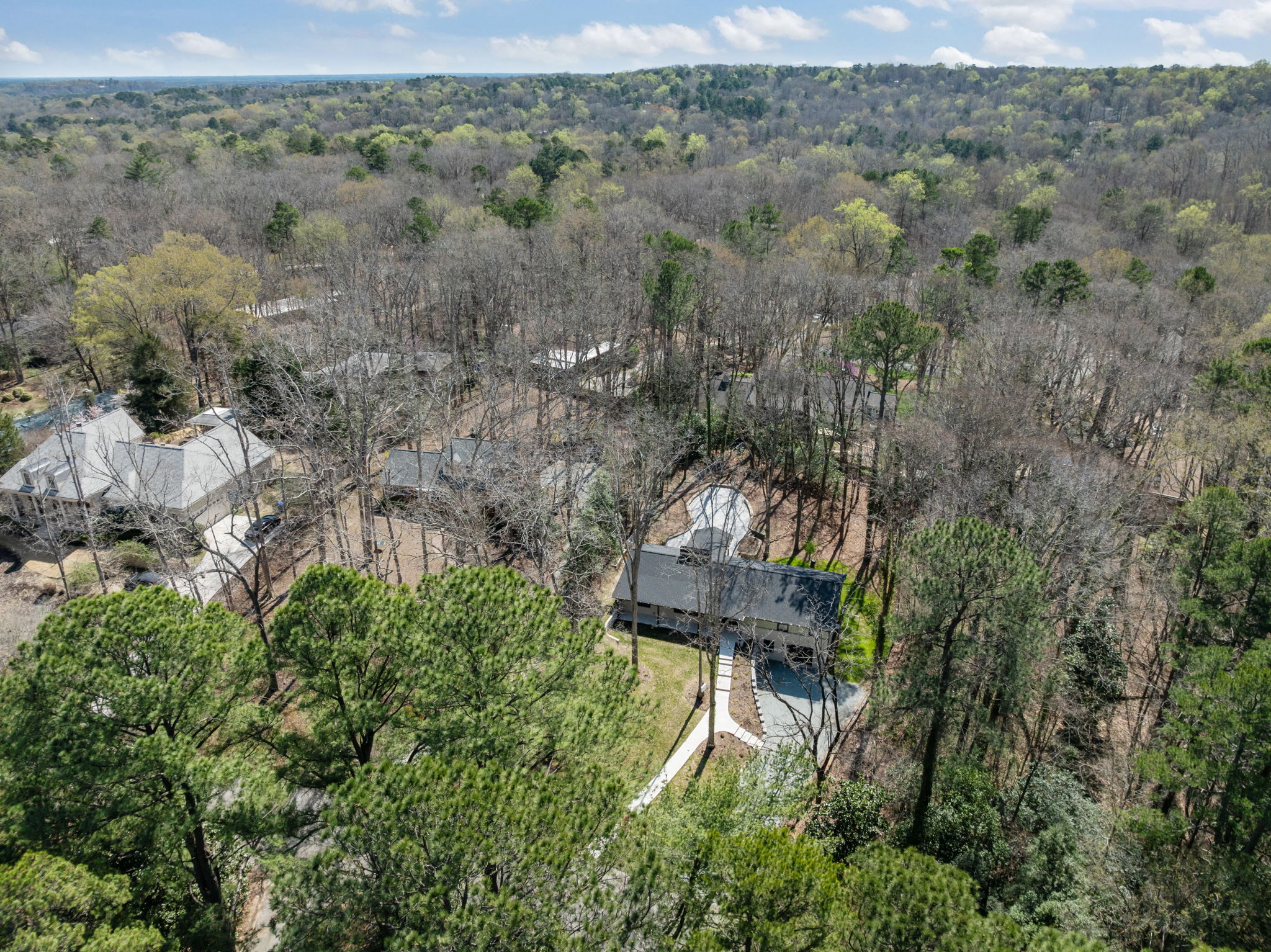 Aerial view of a single-story home surrounded by dense trees with a curved driveway and neighboring houses nearby.