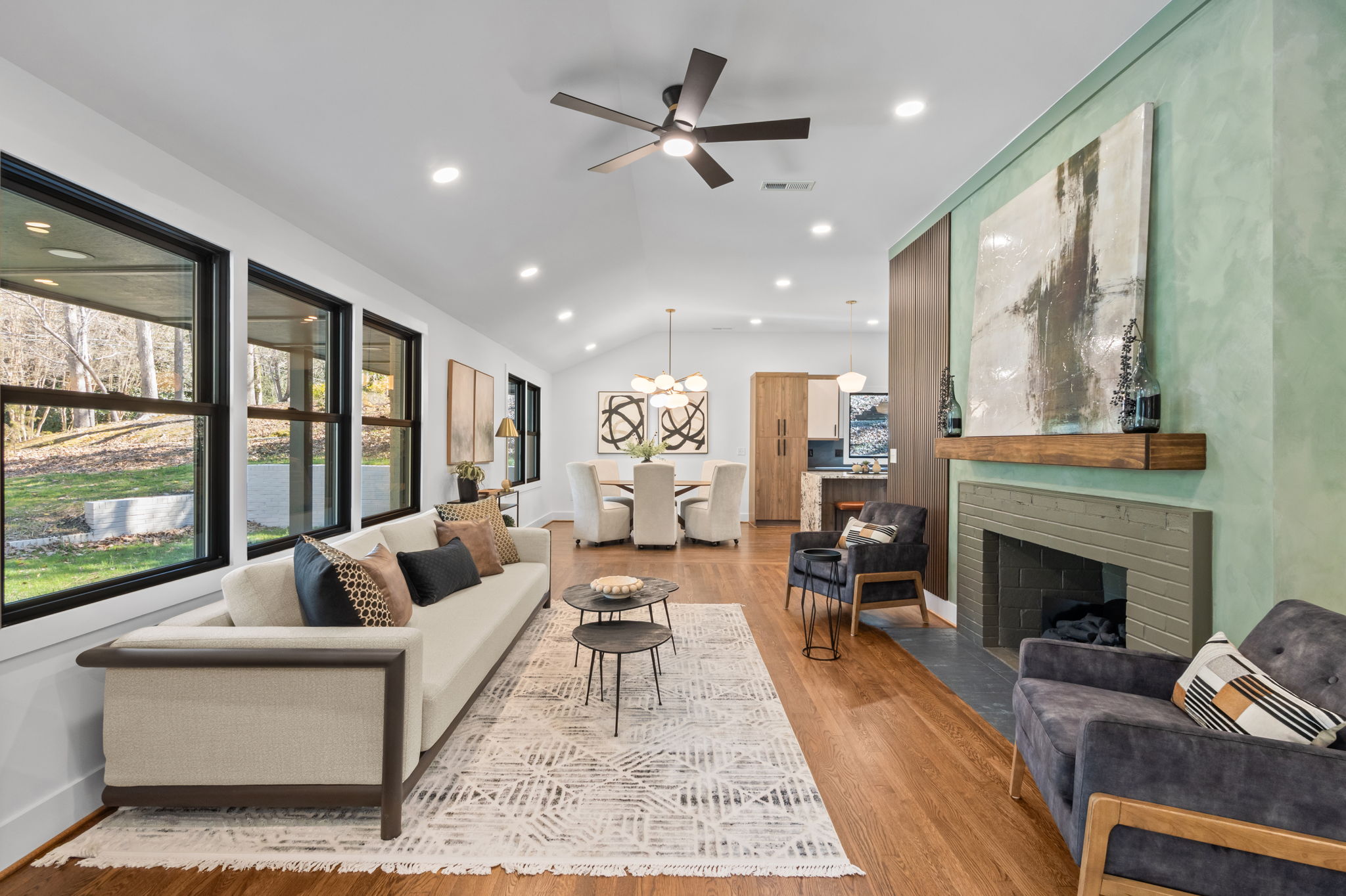 Bright living room with large windows, hardwood floors, fireplace with green accent wall, and seating area leading to a dining space.