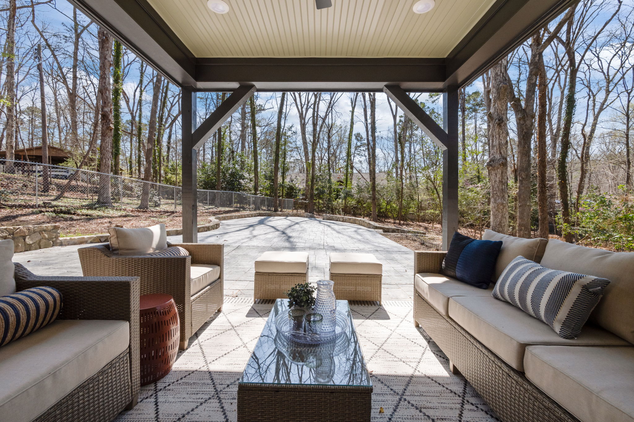 Covered outdoor patio with seating area overlooking a fenced backyard and wooded surroundings.