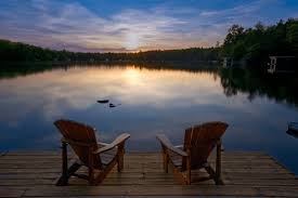 Aerial or dock-level shot of a Maine lake at golden hour — soft reflections, lush pines, one Adirondack chair. No people. Pure feeling.